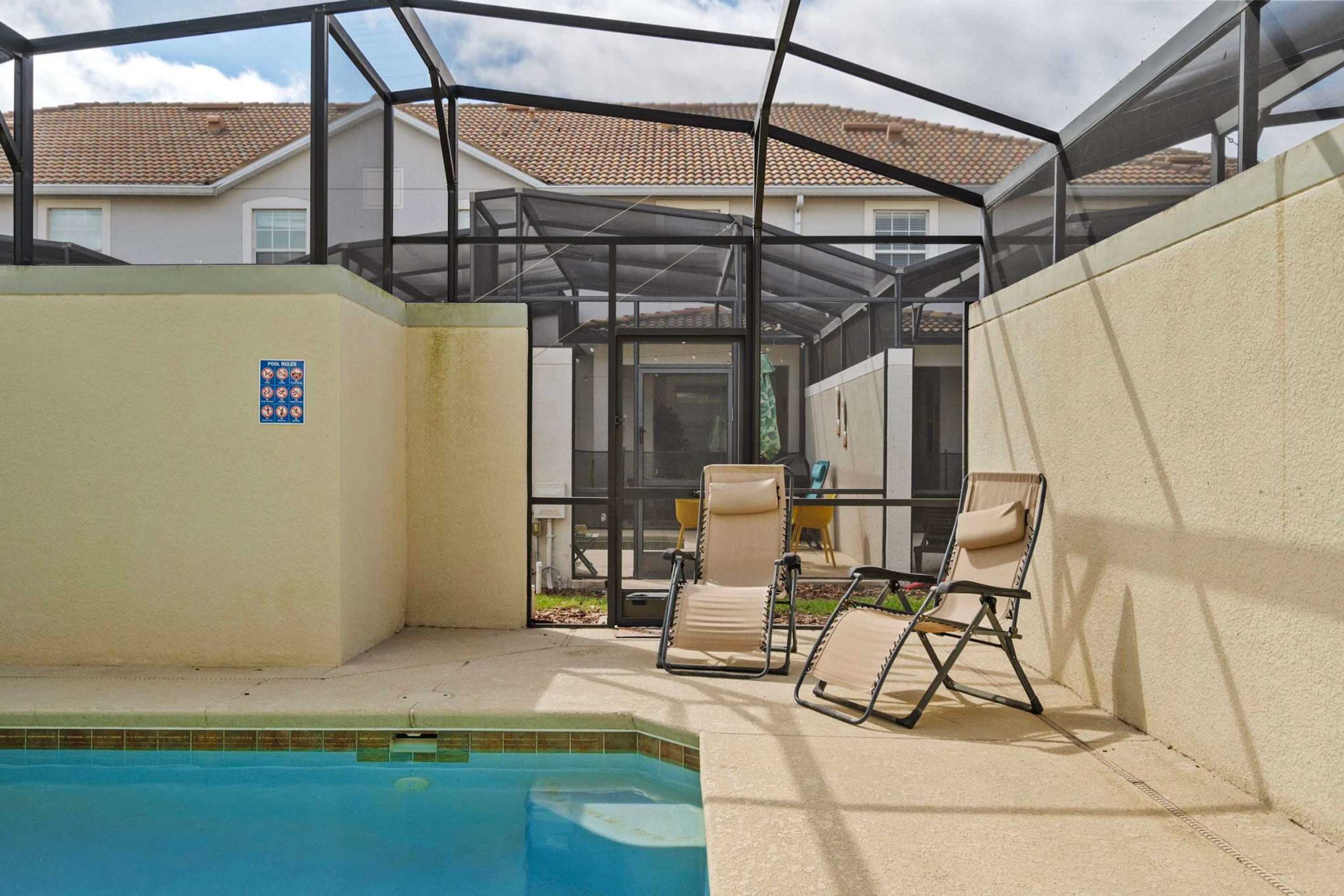 A covered outdoor swimming pool area with two lounge chairs and a patio table.
