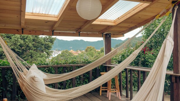 Una hamaca bajo una terraza de madera con tragaluces, con vistas a un paisaje exuberante con montañas.