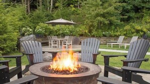 A fire pit surrounded by Adirondack chairs and a table with chairs, set in a garden with trees and a stone wall in the background. - National Apartment (Stowe)