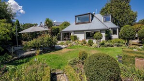 Una casa moderna con techo inclinado, un ventanal grande y un jardín cuidado con diversas plantas y césped.