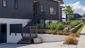 A modern two-story building with a dark exterior, a wooden staircase leading to the entrance, and a landscaped garden with various plants and a small tree. - Peak Retreat - Wanaka Holiday Home (Wanaka)