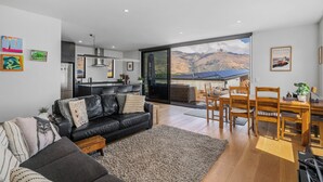 A modern living space with a kitchen, dining area, and a large sliding glass door leading to a deck with a mountain view. - Peak Retreat - Wanaka Holiday Home (Wanaka)