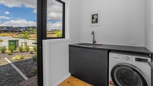A compact kitchen with a built-in washer and dryer, a window with a view of a residential area, and a small countertop. - Peak Retreat - Wanaka Holiday Home (Wanaka)