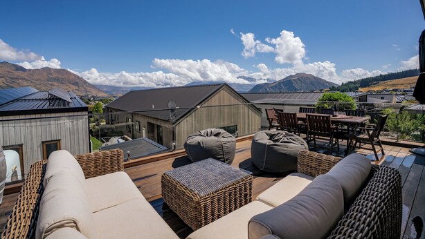 A rooftop terrace with outdoor seating, a view of mountains, and a wooden building in the background. - Peak Retreat - Wanaka Holiday Home (Wanaka)