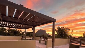 Un espacio al aire libre con bancos y mesas de madera, una estructura de pérgola y una vista de montaña al fondo.