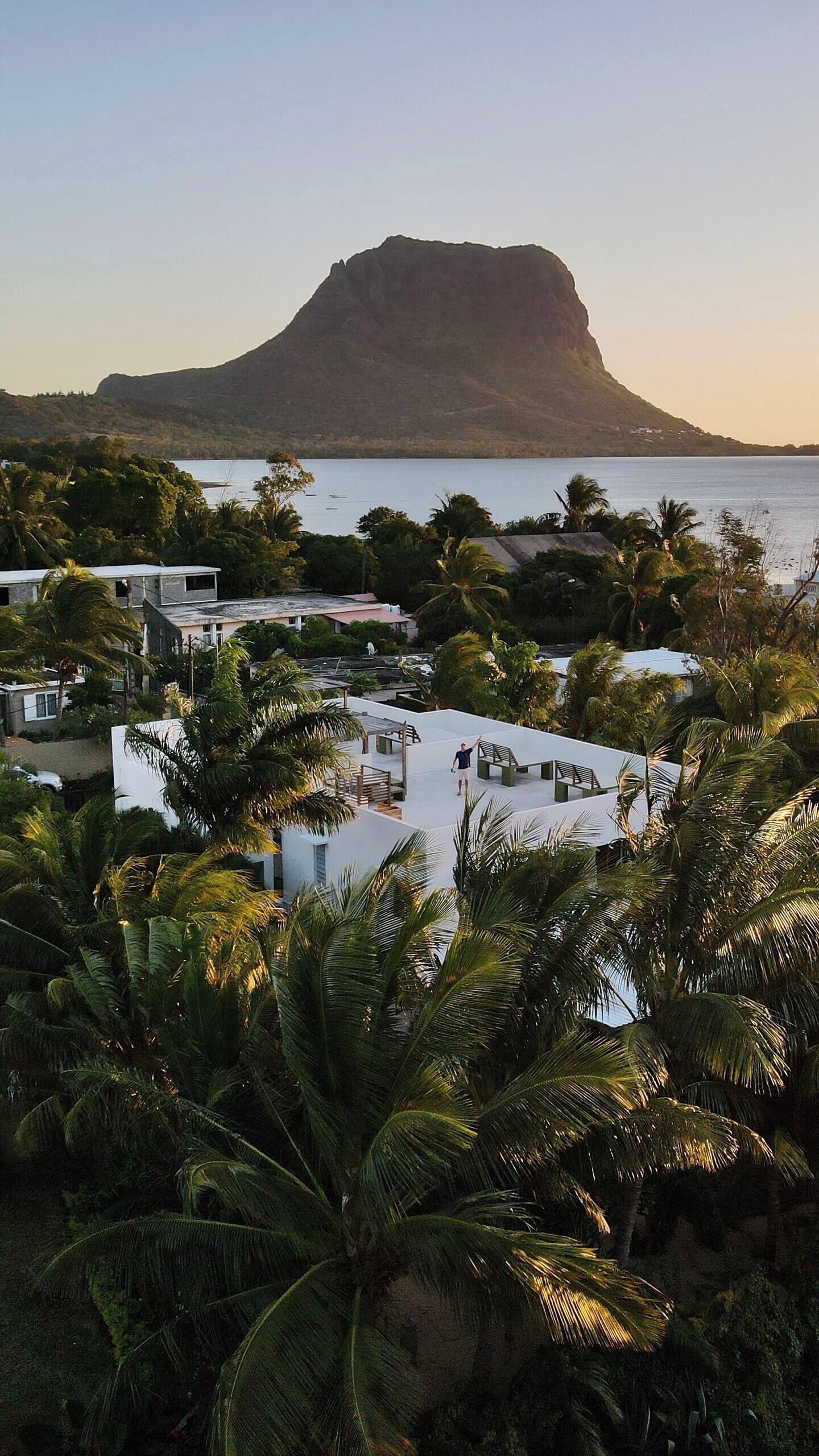 A resort with a pool area surrounded by palm trees, overlooking a body of water and a prominent mountain in the background.