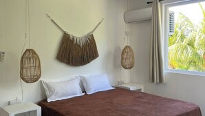 Un dormitorio con una cama, mesitas de noche blancas, un adorno colgante de ratán, una lámpara de mimbre y una ventana con cortinas.