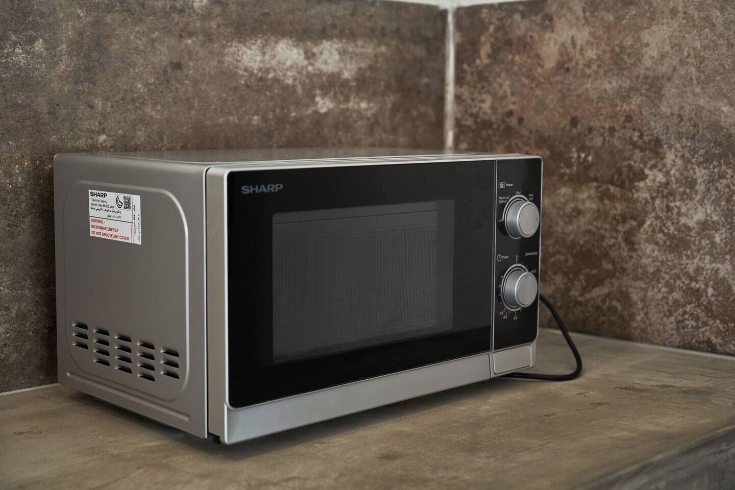 A microwave oven with a warning label, placed on a countertop against a textured wall.