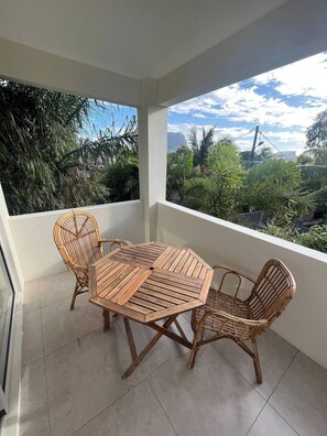 A balcony with a wooden table and wicker chairs, overlooking a garden with various plants and a clear sky.