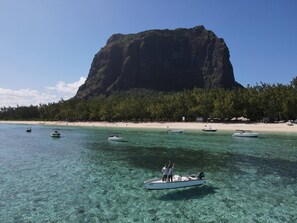 A beach with clear turquoise water, several boats, and a large mountain in the background.