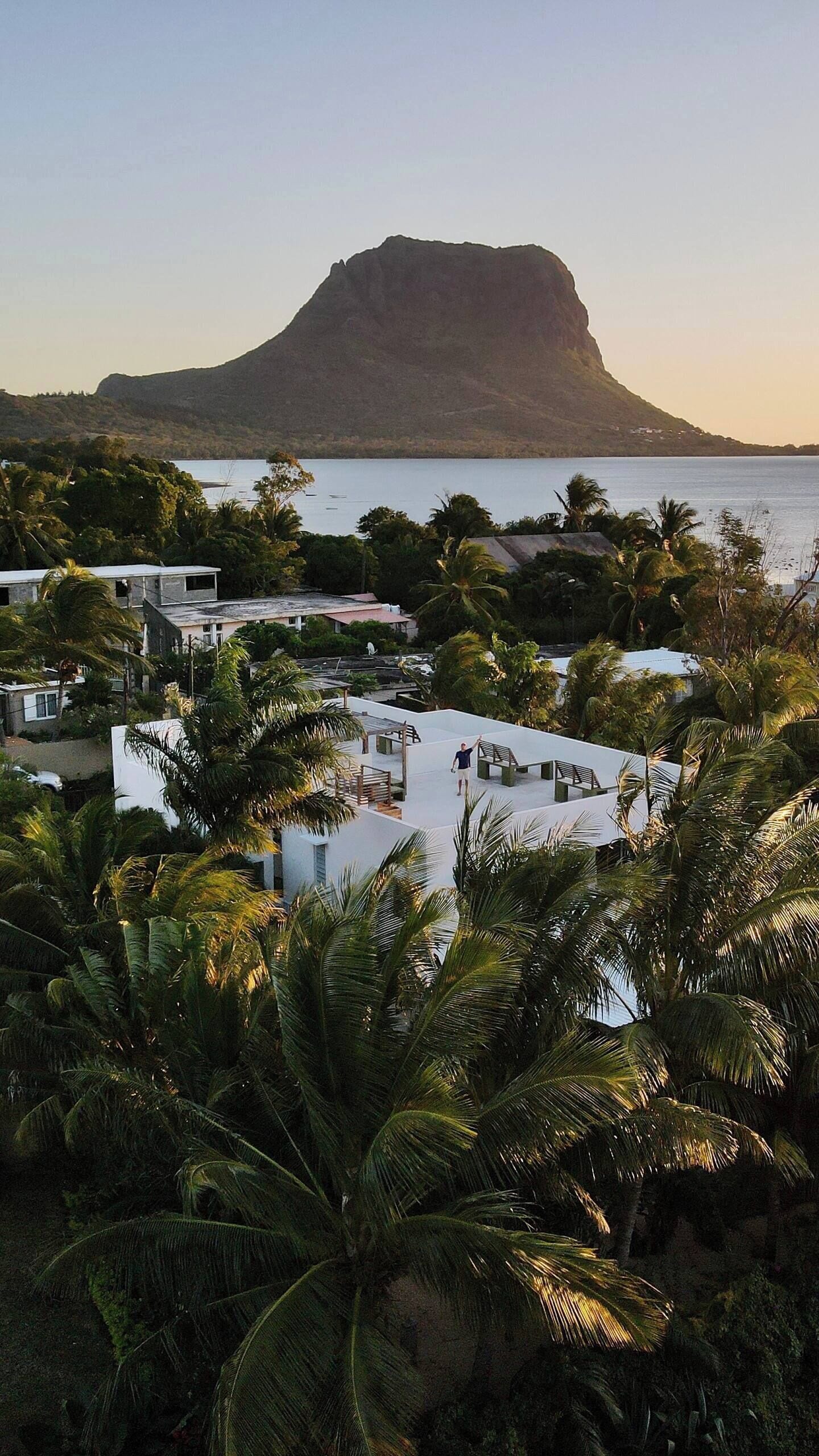 Un complexe hôtelier doté d’une piscine entourée de palmiers, offrant une vue sur une étendue d’eau et sur une montagne imposante en arrière-plan.