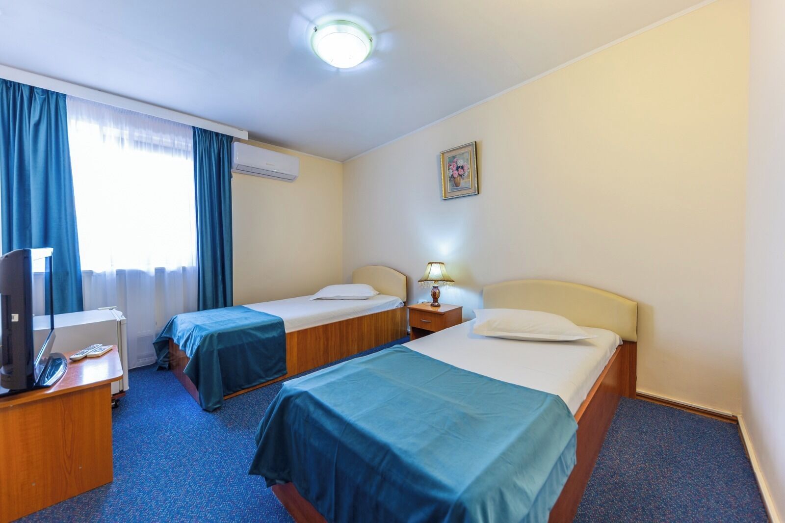 A hotel room with two single beds, a desk with a computer, a TV, and a window with curtains.