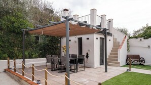 Una casa pequeña y moderna con un patio que cuenta con una mesa, sillas, una pérgola de madera y una escalera que conduce a un nivel superior.