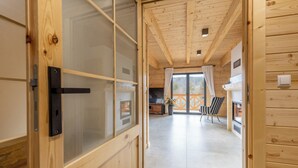 Interior de una cabaña de madera con una puerta corredera, un comedor con mesa y sillas, y un amplio ventanal que ofrece vistas al exterior.