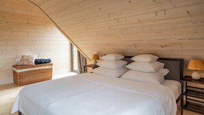 Un dormitorio con techo inclinado, una cama con muchos cojines, una mesita de noche con lámpara y una cesta de mimbre.