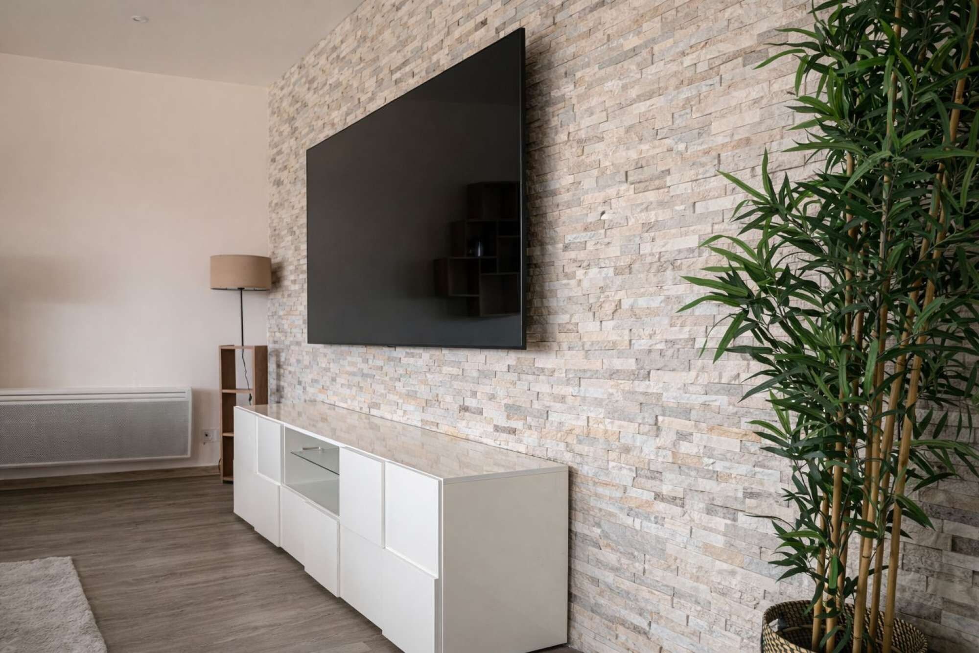Un salon moderne avec un téléviseur à écran plat fixé sur un mur en briques, un meuble blanc en dessous et une plante en bambou sur la droite.