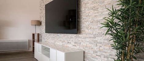 Una sala de estar moderna con un televisor de pantalla plana montado en una pared de ladrillo, un mueble blanco debajo y una planta de bambú a la derecha.