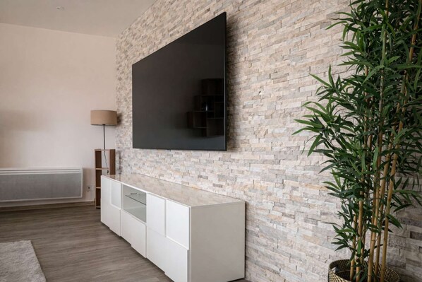 A modern living room with a flat-screen TV mounted on a brick wall, a white cabinet below, and a bamboo plant to the right.