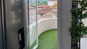 A room with a sliding glass door leading to a balcony with a green artificial turf, a potted plant, and a view of buildings with red roofs. - Les Clés de Laurent – 2-Bedroom Apartment, 70 m², Large Balcony, Downtown (Graulhet)
