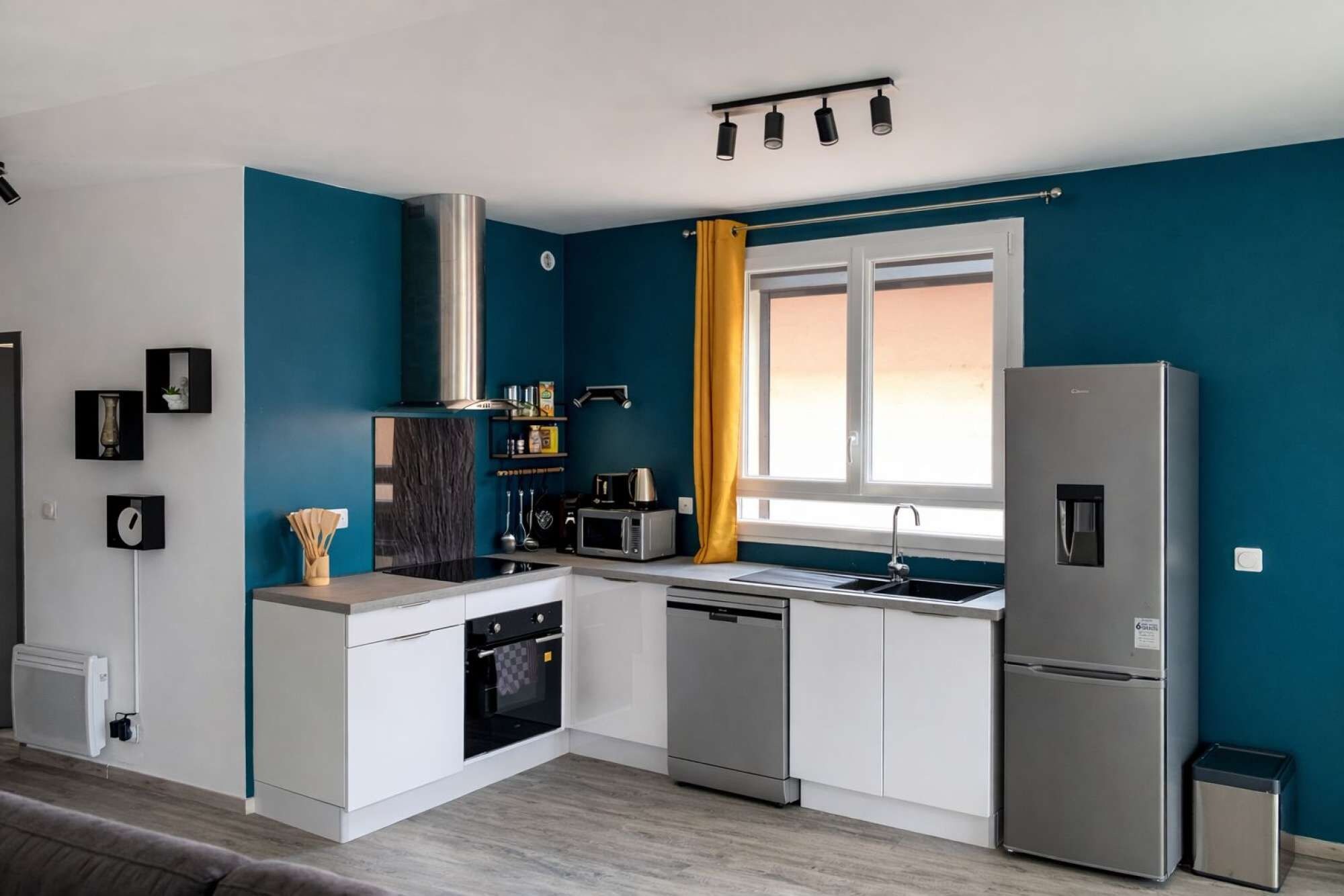 Une cuisine moderne avec des meubles blancs, des appareils en acier inoxydable et une fenêtre ornée de rideaux jaunes.