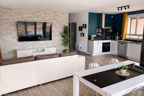 A modern living room with a flat-screen TV, a white sofa, and a dining area with a black table. - Les Clés de Laurent – 2-room apartment, 70 m², large balcony, downtown (Graulhet)
