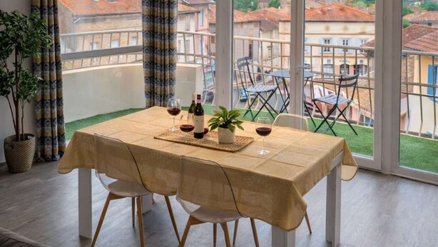 A dining area with a table set for two, a potted plant, and a view of the cityscape through large windows. - Les Clés de Laurent – 2-Bedroom Apartment, 70 m², Large Balcony, Downtown (Graulhet)