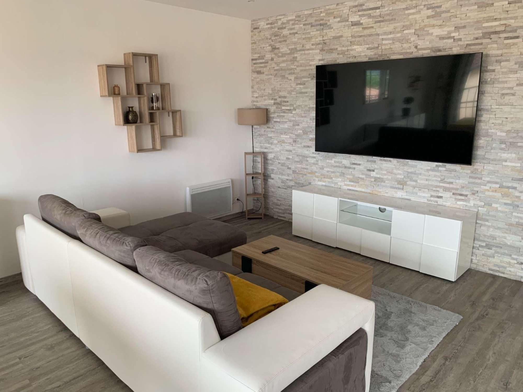 A modern living room with a sectional sofa, a flat-screen TV mounted on a stone-textured wall, a wooden TV stand, and a floor lamp.