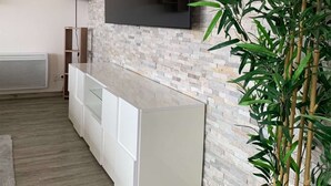 A modern living room with a flat-screen TV mounted on a textured wall, a white console table, a floor lamp, and a large bamboo plant. - Les Clés de Laurent – 2-Bedroom Apartment, 70 m², Large Balcony, Downtown (Graulhet)