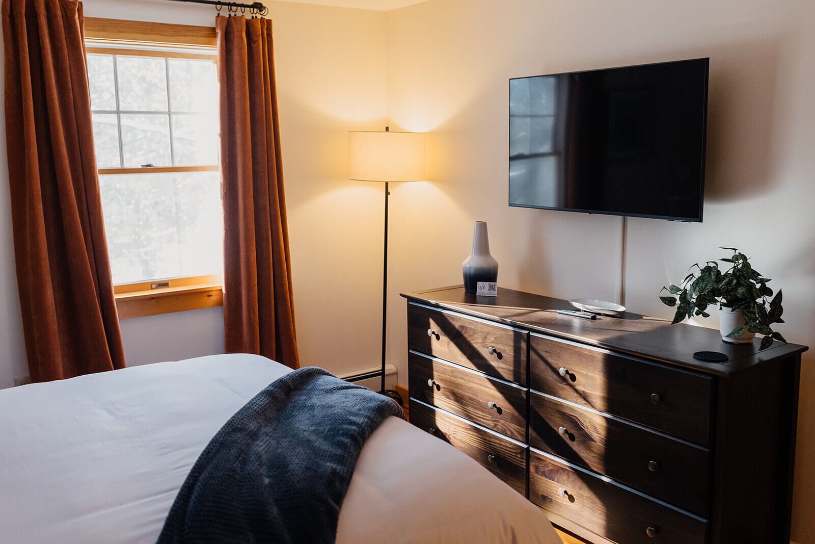 Quarto de hotel com cama, penteadeira, televisão, luminária e janela com cortinas.