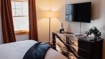 Quarto de hotel com cama, penteadeira, televisão, luminária e janela com cortinas.