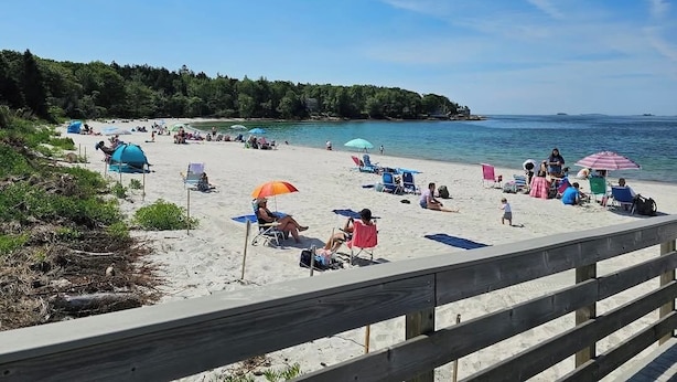 A beach with people relaxing under umbrellas, a wooden fence in the foreground, and a forested area in the background. - Pemaquid Escape | Curated Comfort + Hot Tub (Walpole)