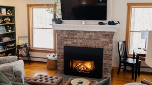 A living room with a brick fireplace, a flat-screen TV mounted above it, and a bookshelf filled with books and decorative items. - Pemaquid Escape | Curated Comfort + Hot Tub (Walpole)
