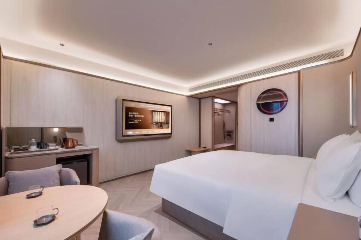A modern hotel room with a bed, a desk with a chair, a television, and a round mirror.