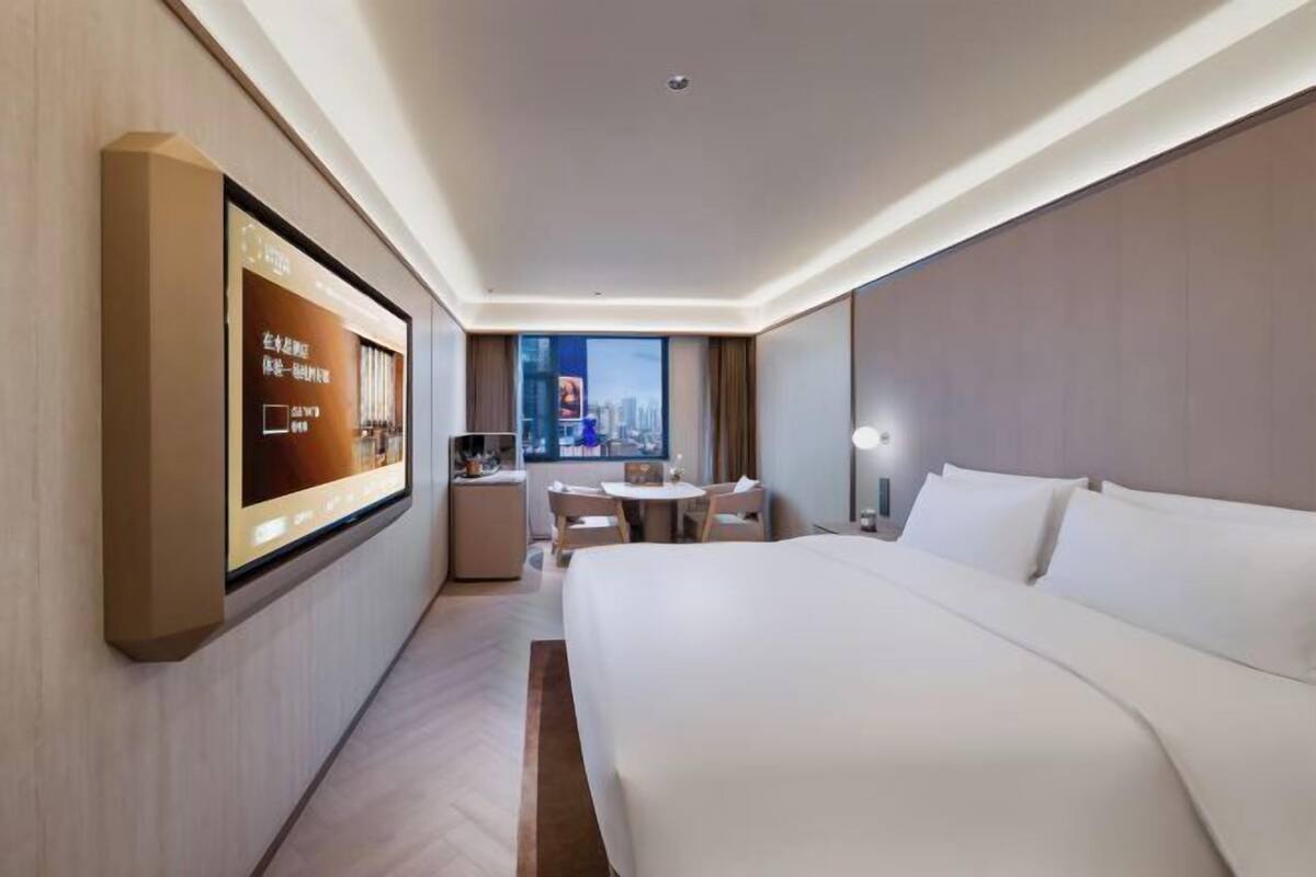 A hotel room with a large bed, a desk with a chair, a flat-screen TV mounted on the wall, and a cityscape view.