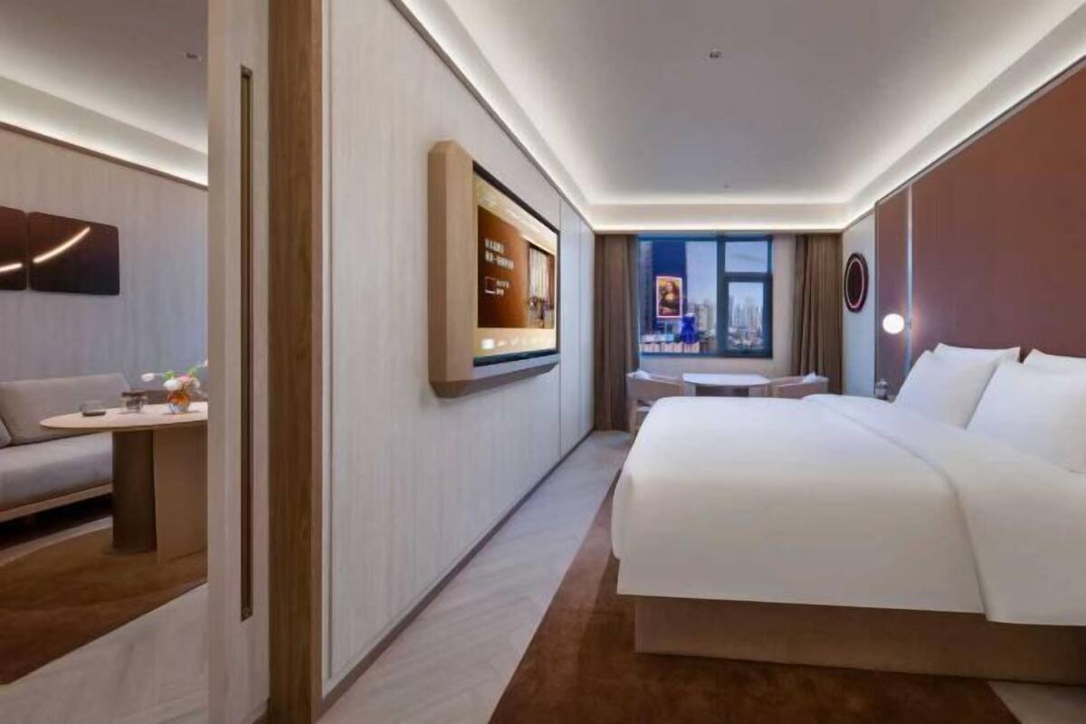 A modern hotel room with a large bed, a seating area with a table, a TV mounted on the wall, and a window with a city view.