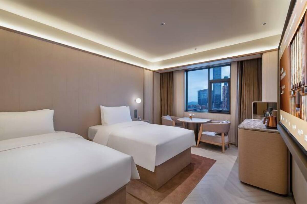 A hotel room with two beds, a small desk, a chair, and a large window with a city view.