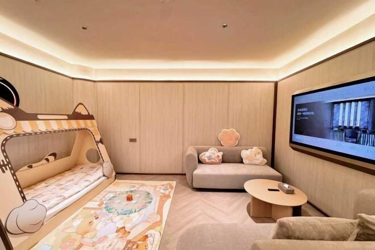 A modern, well-lit room with a large TV screen, a sofa, a small round table, and a children's play area with a mat and toys.