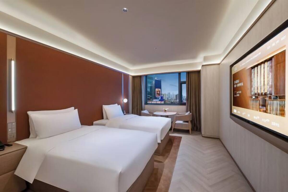 A hotel room with two beds, a desk, a chair, a TV screen, and a window with a city view.
