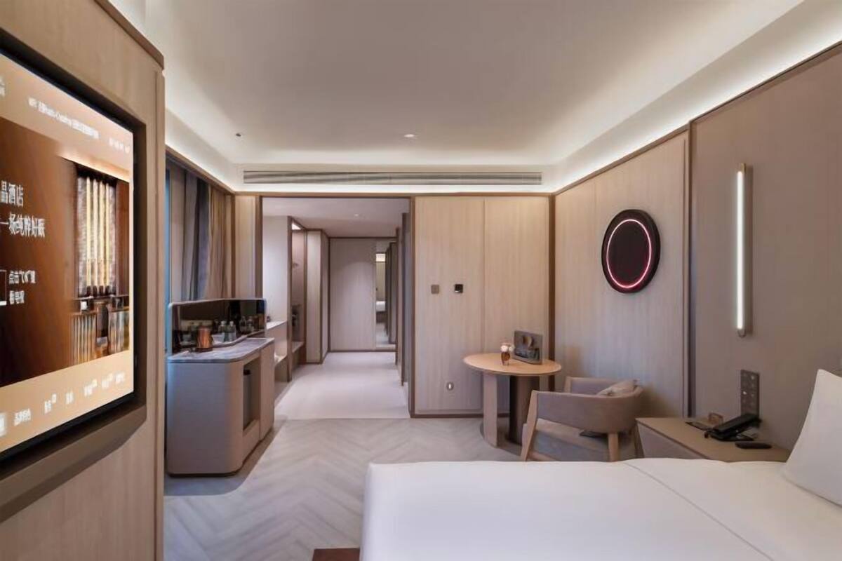 A hotel room with a bed, bedside table, a small round table with a chair, a TV mounted on the wall, and a minibar.