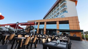 An outdoor dining area with tables and chairs, a building with multiple floors and balconies, and a clear blue sky. - Throne Beach Resort & Spa (Manavgat)