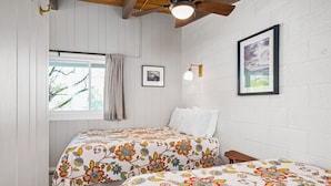 Un dormitorio con dos camas, paredes blancas, ventilador de techo, ventana con cortinas e imagen enmarcada en la pared.