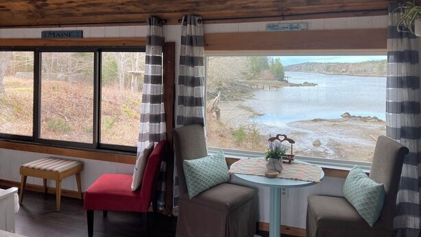 A cabin interior with a large window overlooking a river, wooden ceiling, and a small table with a decorative item. - Enjoyable 1-bedroom rustic cottage in Southport. (Southport)