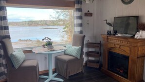 A cozy room with a view of a lake, featuring two chairs, a small table, a fireplace, and a television. - Enjoyable 1-bedroom rustic cottage in Southport. (Southport)