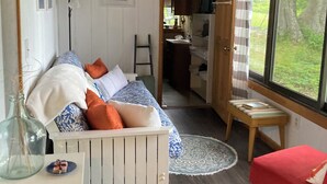 A cozy room with a wooden ceiling, a white sofa with patterned cushions, a small wooden table, and a view of greenery outside. - Enjoyable 1-bedroom rustic cottage in Southport. (Southport)