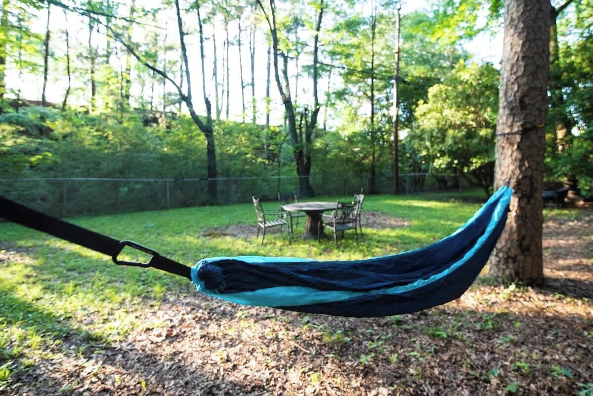 A hammock suspended between trees in a wooded area with a table and chairs in the background.