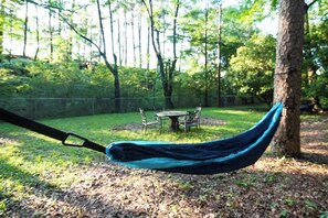 A hammock suspended between trees in a wooded area with a table and chairs in the background. - Spacious, stylish home with fenced yard! (Little Rock)