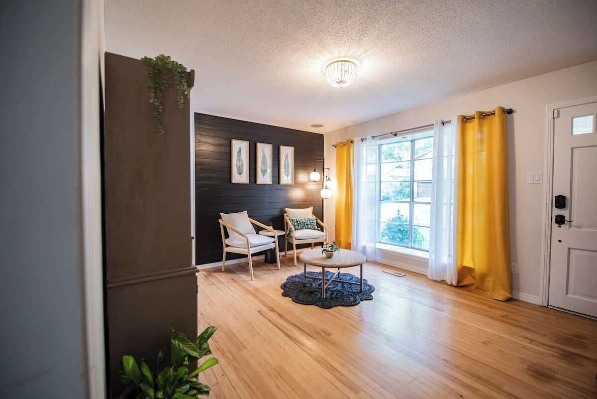 A room with a wooden floor, two chairs, a small table, a ceiling light, and a window with yellow curtains.