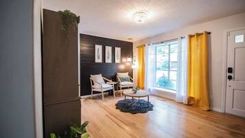 A room with a wooden floor, two chairs, a small table, a ceiling light, and a window with yellow curtains.