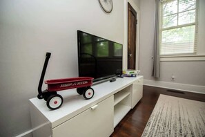 Une pièce avec un téléviseur posé sur un meuble blanc, un petit chariot jouet rouge et une fenêtre avec des stores.