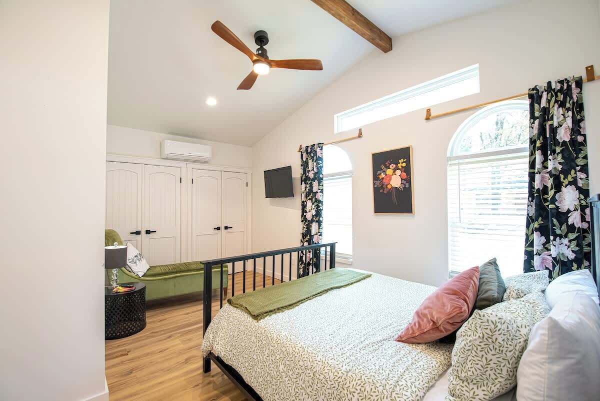 A bedroom with a bed, a ceiling fan, a window with curtains, and a wall-mounted TV.